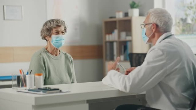 中年家庭医生与老年女病人在门诊会诊期间交谈。两人都戴着口罩。内科医生穿着白大褂，坐在医院办公室的桌子