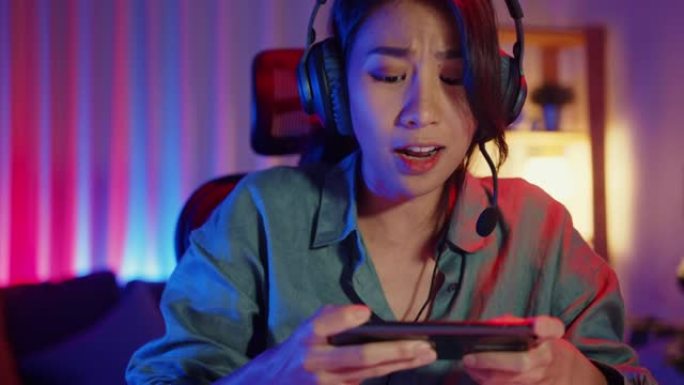 快乐亚洲女孩游戏玩家戴着耳机比赛在线玩视频游戏，在家里的客厅用智能手机彩色霓虹灯。