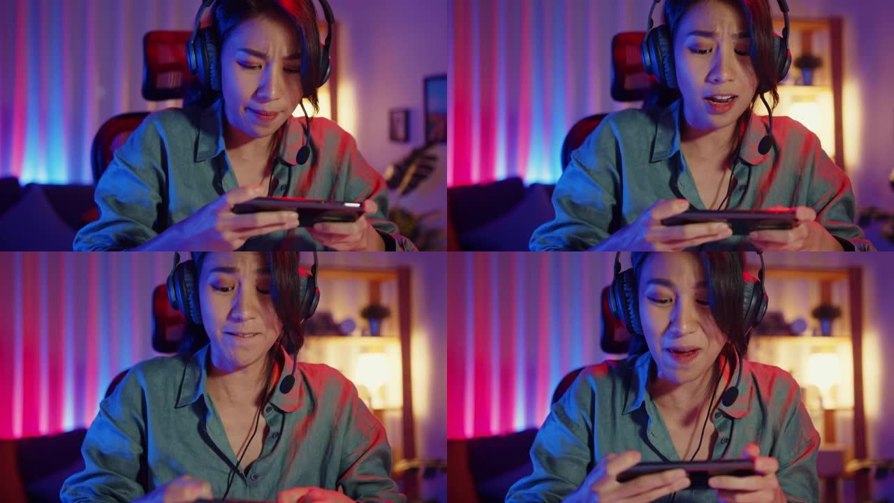 快乐亚洲女孩游戏玩家戴着耳机比赛在线玩视频游戏，在家里的客厅用智能手机彩色霓虹灯。