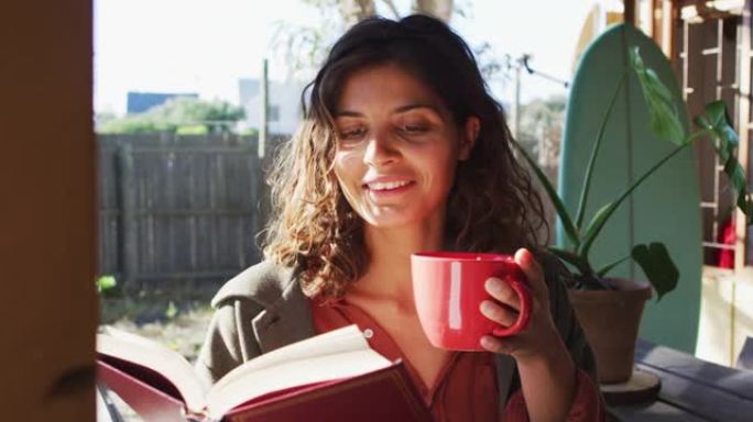 快乐的混血女人坐在阳光明媚的小屋厨房喝茶看书
