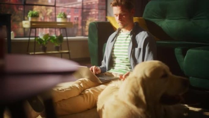 英俊的年轻人使用笔记本电脑，在家客厅工作。男性坐在地板上，使用电脑，金毛狗坐在他旁边，想玩耍和被爱抚
