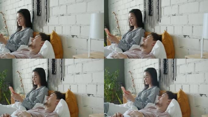 快乐的亚洲女士玩智能手机，男朋友在家睡觉