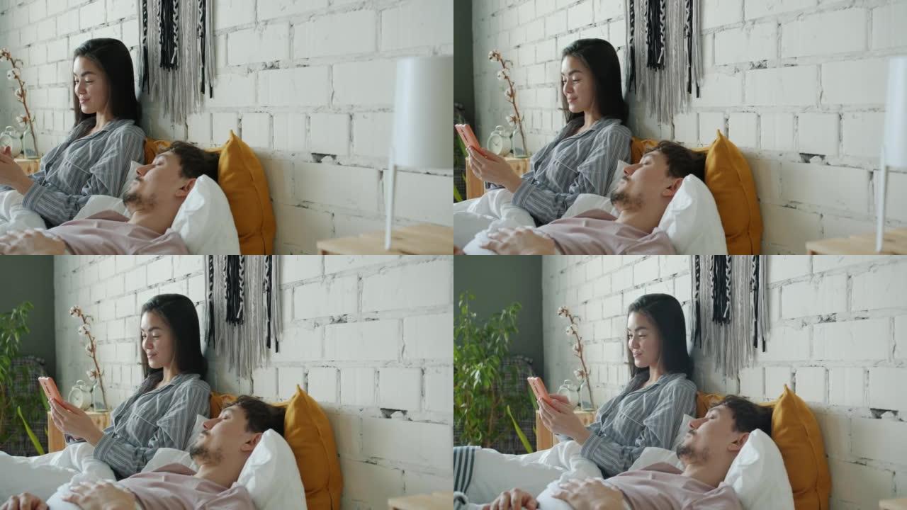 快乐的亚洲女士玩智能手机，男朋友在家睡觉