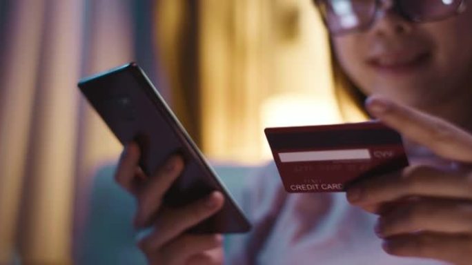 亚洲女性使用智能手机在客厅沙发上订购带信用卡的网上购物产品