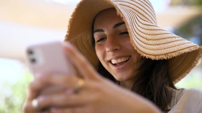 一位戴着草帽的快乐无忧无虑的微笑年轻女性游客在阳光明媚的日子里，正在使用智能手机发送消息，拨打电话或