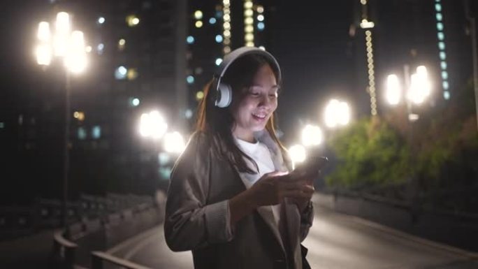 下班后，在城市晚上使用带耳机的智能手机行走的亚洲妇女