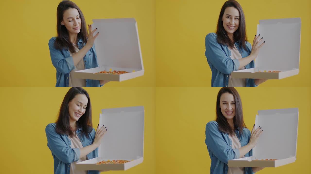 年轻女子打开纸板箱看着披萨并在黄色背景上微笑的肖像