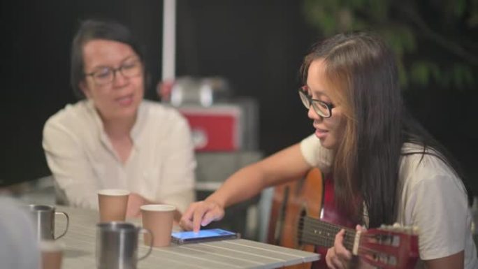 亚洲中国女儿从智能手机中选择歌曲阅读歌词哼唱吉他，与母亲和朋友一起在晚上的花园聚会露营帐篷中享受