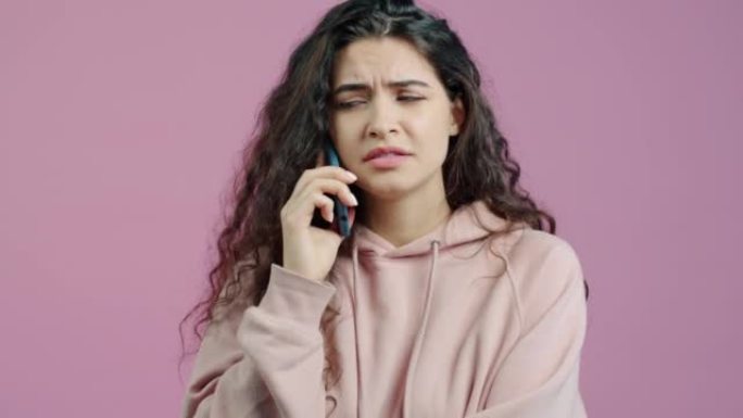 愤怒的年轻女子站在粉红色的背景上在手机上讲话