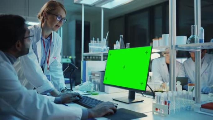 女研究科学家在带有绿屏模型的台式计算机旁边与生物工程师进行了对话。他们在现代科学实验室中查看计算机模