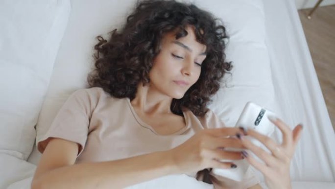穿着睡衣的开朗女孩在家床上醒来触摸智能手机屏幕然后伸展手臂