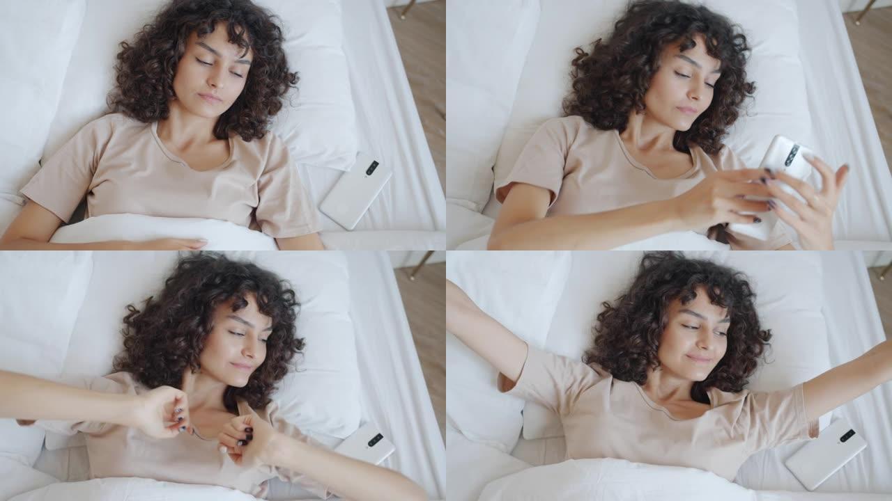 穿着睡衣的开朗女孩在家床上醒来触摸智能手机屏幕然后伸展手臂