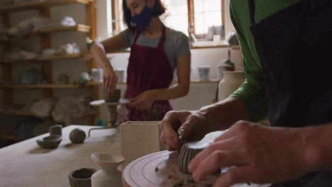 男性陶工戴着口罩和围裙，使用丝带工具在陶器工作室的锅上进行设计