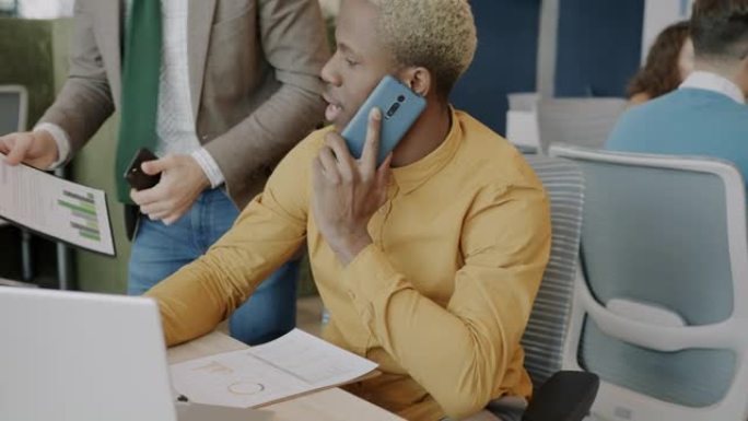 忙碌的非洲裔美国员工使用笔记本电脑在手机上讲话并帮助同事处理文件