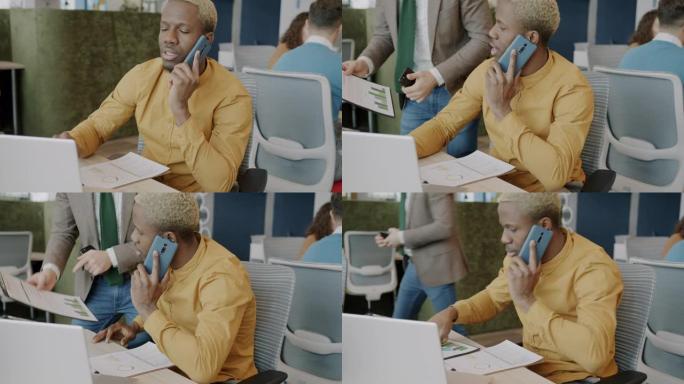 忙碌的非洲裔美国员工使用笔记本电脑在手机上讲话并帮助同事处理文件