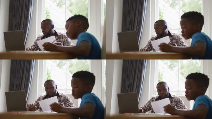 非裔美国人在家里使用笔记本电脑工作，与儿子一起坐在桌子旁做学校工作