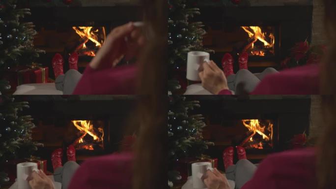 年轻女子喝茶并在圣诞树旁看壁炉的后视图