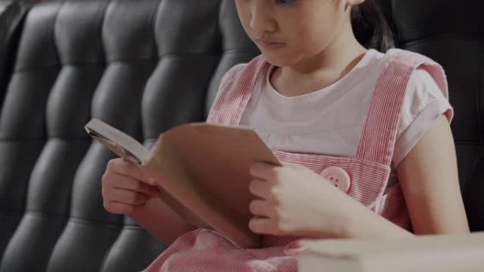 亚洲小女孩坐在沙发上看书，呆在家里。她翻开学习书的一页。学习教育书籍的女孩。