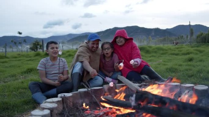 美丽的农民家庭在田间工作后享受篝火，烤棉花糖说话和微笑
