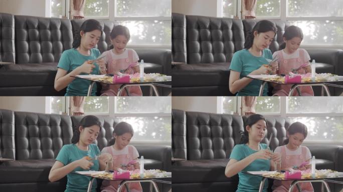 两个亚洲兄弟姐妹在家里的同时在客厅剪纸并制作手工艺品。姐姐用彩纸，剪刀和胶水在纸上创作艺术作品，形成