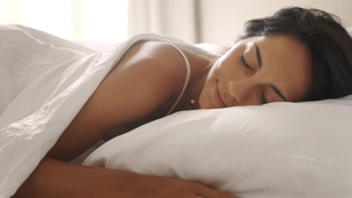 一名年轻黑发女子的真实特写镜头正在卧室舒适的床上，裹着温暖的羽绒被毯子安然入睡。舒适、放松、睡眠、健