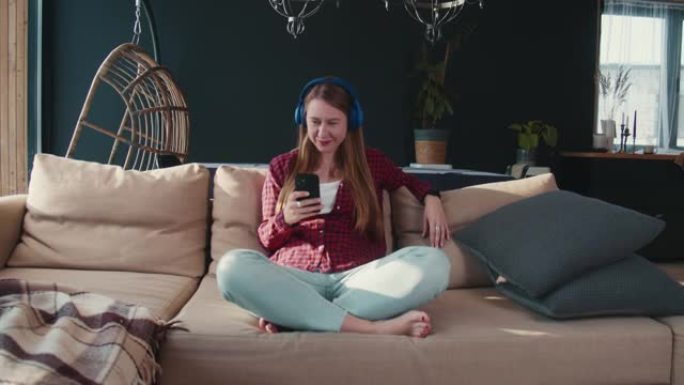 快乐美丽的高加索千禧一代金发女子戴着耳机向音乐点头，使用智能手机浏览新闻。