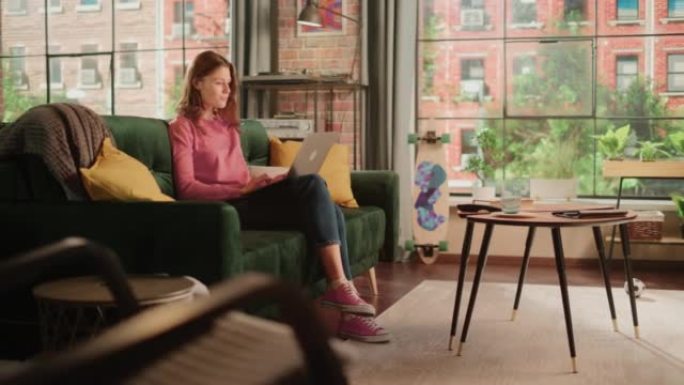 年轻漂亮的女性坐在客厅的沙发上，在家用笔记本电脑工作。有创造力的女人检查社交媒体，浏览互联网。从大窗