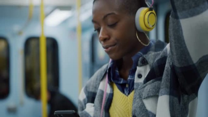 微笑的黑人妇女在乘地铁旅行时使用智能手机和耳机听音乐。年轻女性使用公共交通工具回家，浏览社交媒体，在
