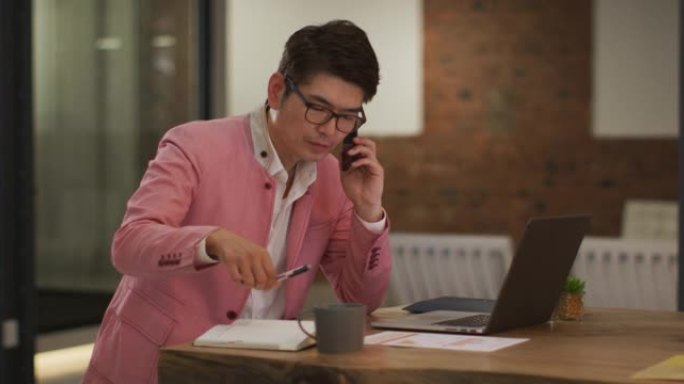 穿着时髦的亚洲商人坐在办公桌前，用笔记本电脑做笔记