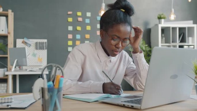 非裔美国女商人使用笔记本电脑在笔记本上做笔记，看起来很累又不开心