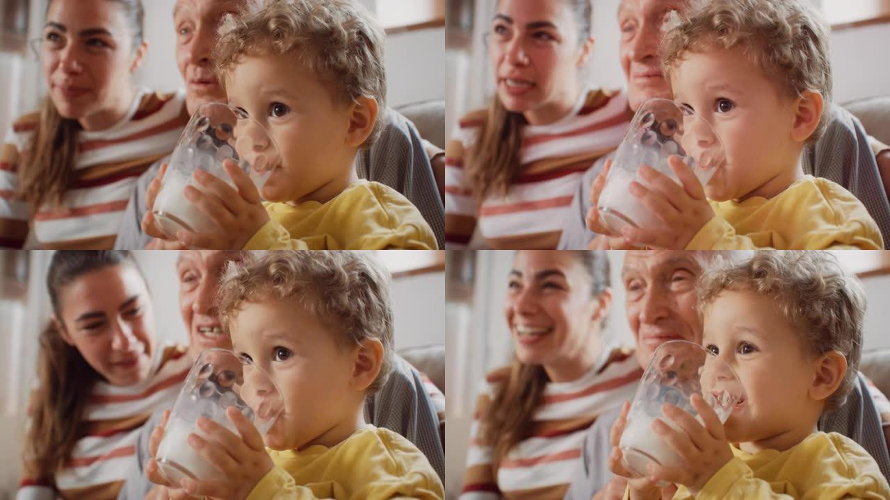 孩子在客厅喝牛奶时与家人一起看电视的肖像。他的母亲和祖父和他说话。可爱的蹒跚学步的孩子为他的成长和健