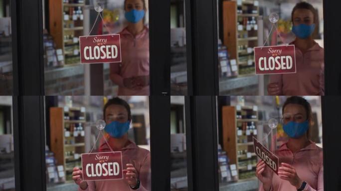 戴口罩的高加索女店主在运动商店的橱窗中关闭以打开标志