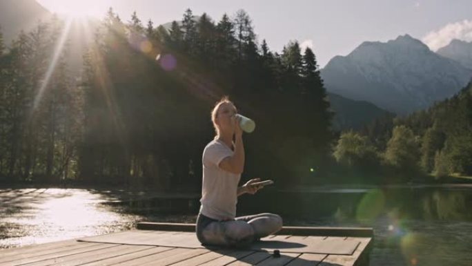 金色头发的成年女性，坐在湖前的瑜伽马特上，休息一下训练，喝酒和使用手机