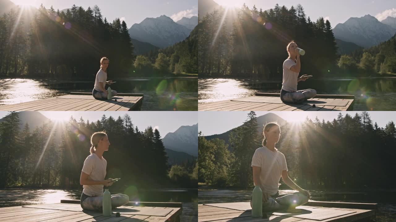 金色头发的成年女性，坐在湖前的瑜伽马特上，休息一下训练，喝酒和使用手机