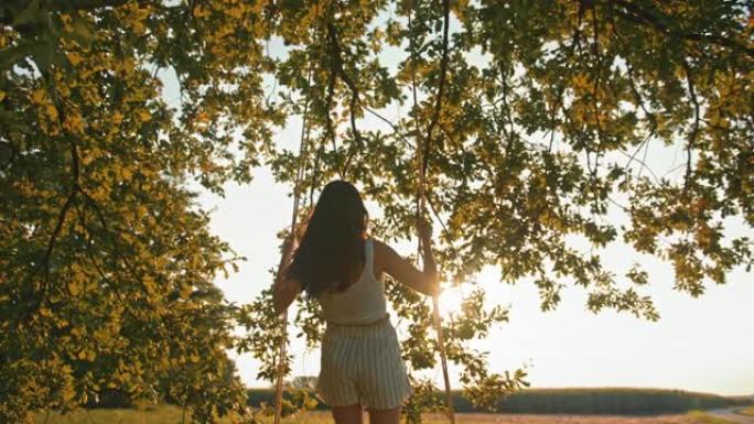 慢动作，手持镜头拍摄一个年轻的赤脚女孩，站在阳光充足的草地上悬挂在大树枝上的绳索秋千上
