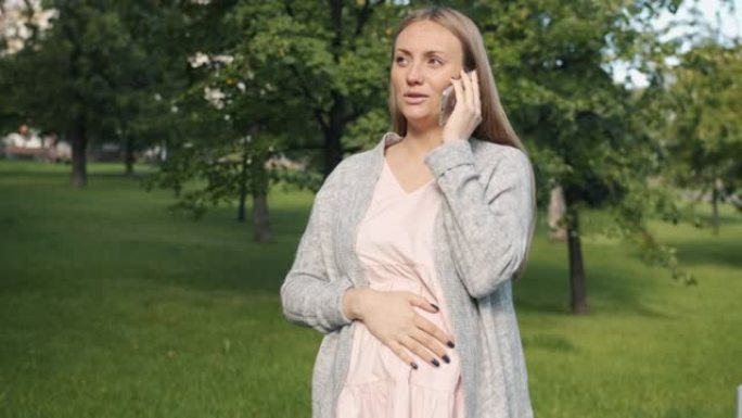 快乐的孕妇在夏日站在城市公园的手机上讲话