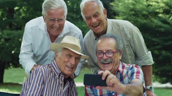 一群老人在公园里享受户外天气。老年男性朋友在智能手机上拍照时开玩笑地做鬼脸。积极活泼的老年人使用技术