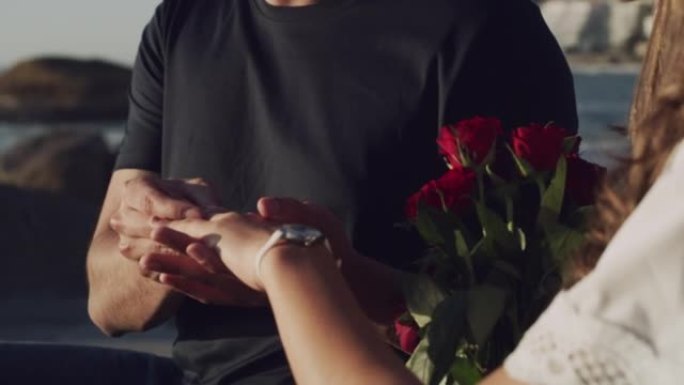 一对浪漫的夫妇在海滩求婚后订婚。年轻女子拿着一束红玫瑰，在未婚夫的手指上戴了一枚钻戒后，用爱意的手势