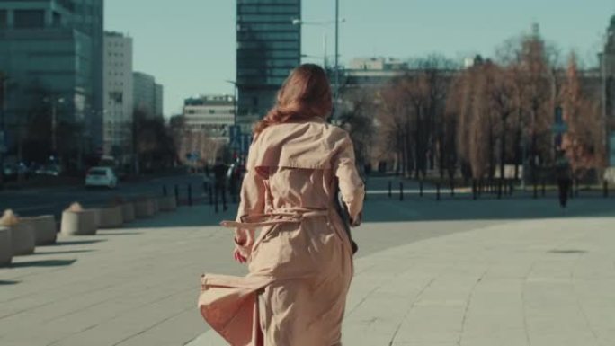 镜头跟随美丽的女人穿着时尚的浅米色秋衣独自走在阳光明媚的大城市广场慢动作。