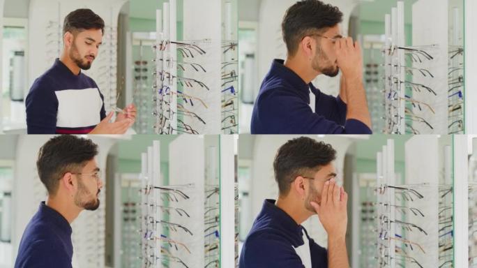 在验光诊所选择眼镜的年轻人。患者尝试并测试验光师处方镜片的眼镜架。男人用镜子欣赏眼镜师戴的新视觉的反