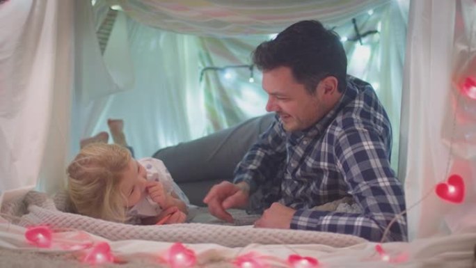 父亲和年幼的女儿在家里孩子卧室的自制营地里用彩色书