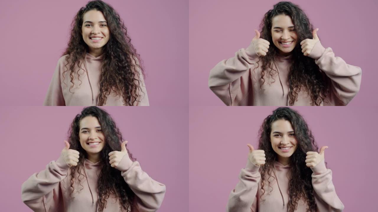 快乐的年轻女子表现出竖起大拇指的手势，并在粉红色背景下微笑