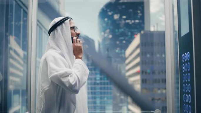 成功的穆斯林商人在传统的白色坎杜拉骑着玻璃电梯到现代商务中心的办公室。男子在电梯里打电话。沙特、阿联