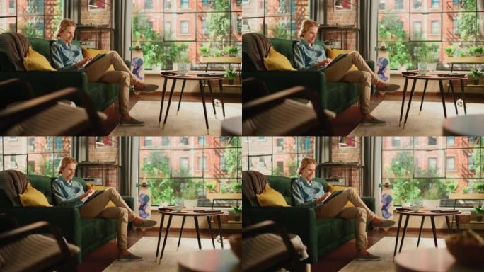 年轻英俊的成年男子坐在阁楼公寓的沙发上，在家用平板电脑工作。创意男性查看社交媒体，浏览互联网。从大窗