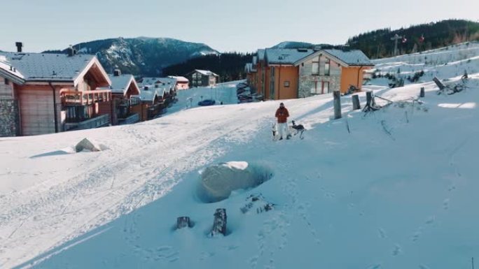 无人机在美丽的冬季度假村建筑附近低空飞行，人与狗在白雪覆盖的晨山景观中。