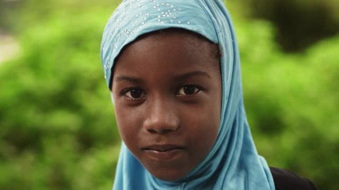 一个害羞的地道的非洲小女孩对着镜头微笑的特写肖像，背景是模糊的绿色。代表未来、希望和接受的黑人女性孩