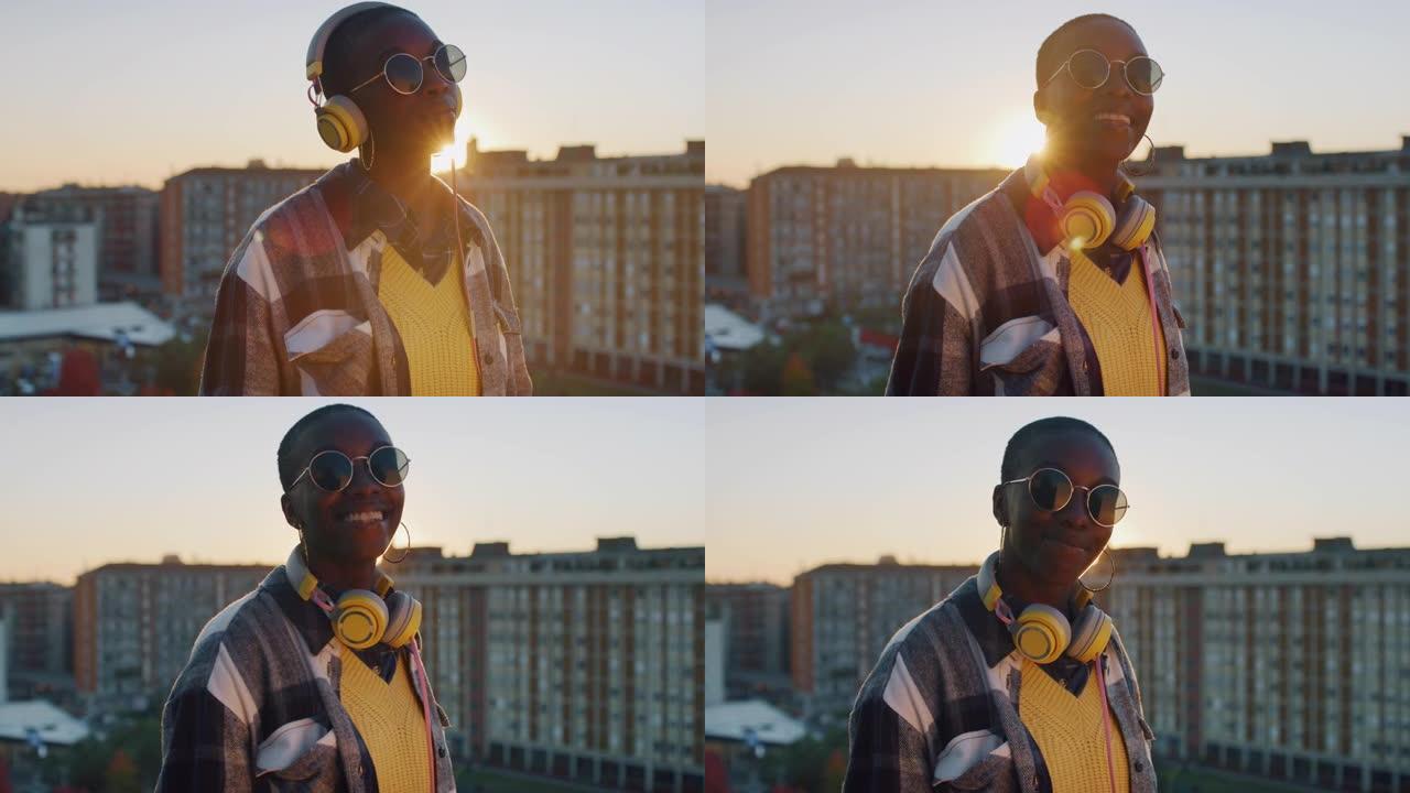 快乐的年轻黑人妇女在屋顶上使用耳机，然后摘下耳机，微笑着看着相机。日落时欣赏音乐和城市景观的非洲裔美