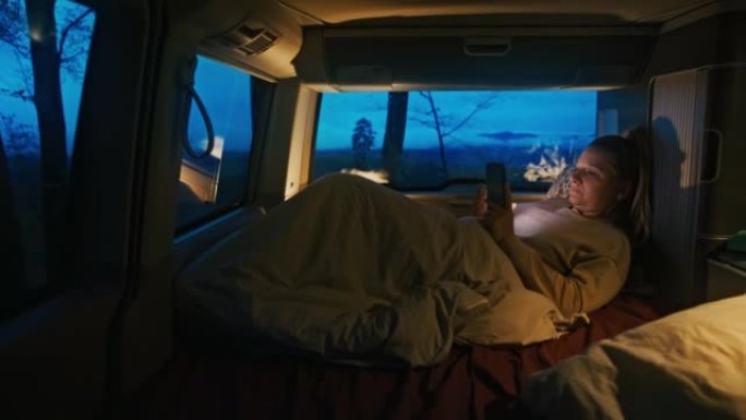 金发碧眼的女人躺在毯子下的货车里，晚上用智能手机