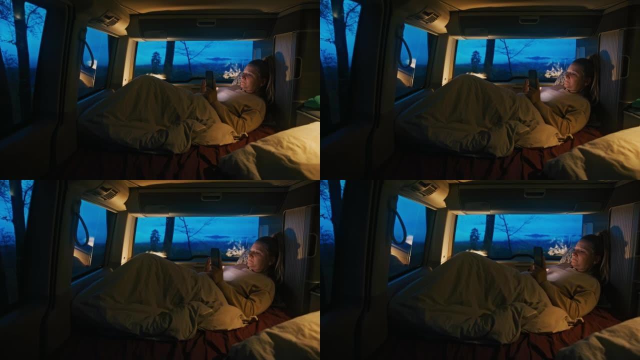 金发碧眼的女人躺在毯子下的货车里，晚上用智能手机