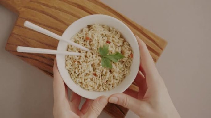 双手在米色背景上放一碗筷子方便面的俯视图